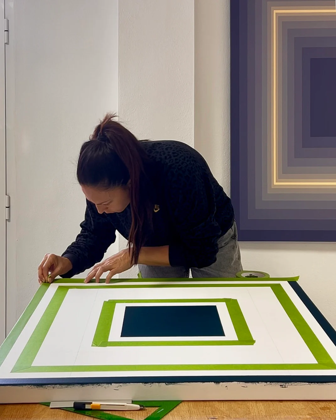 Martina Berti carefully applies green masking tape to a geometric canvas in progress, creating precise square shapes. She is working at a wooden table in a bright studio, with a completed artwork of layered colored squares hanging on the wall behind her.